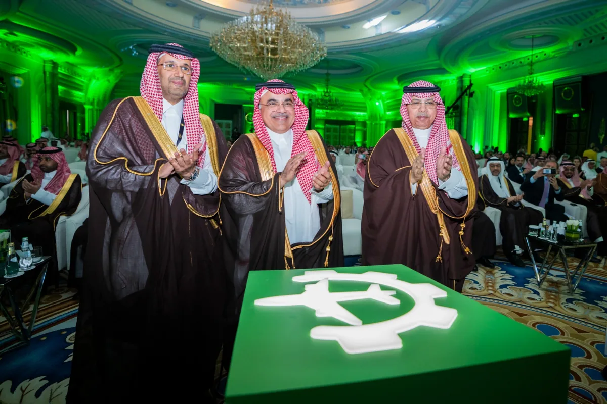 Three officials applaud at the green reveal plinth, its emblem lit from within, at the ministerial launch ceremony.